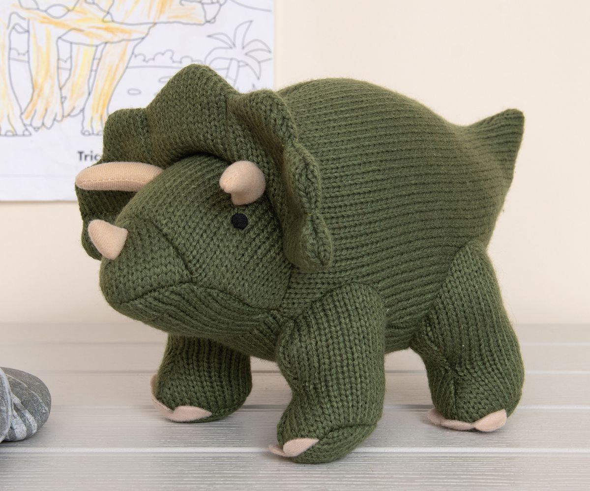 Knitted moss green triceratops dinosaur soft toy on white floorboards with drawing on the wall behind